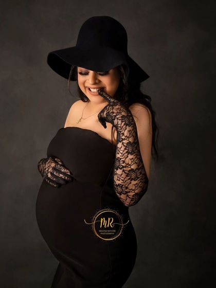A cute and playful close-up portrait. The mother-to-be, dressed in a chic black hat and gloves, gives a joyful smile to the camera.