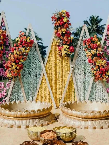 A modern Haldi setup featuring triangular backdrops decorated with marigolds and other colorful flowers. The seating area includes two large brass urlis for the ceremony, blending geometric shapes with traditional elements.