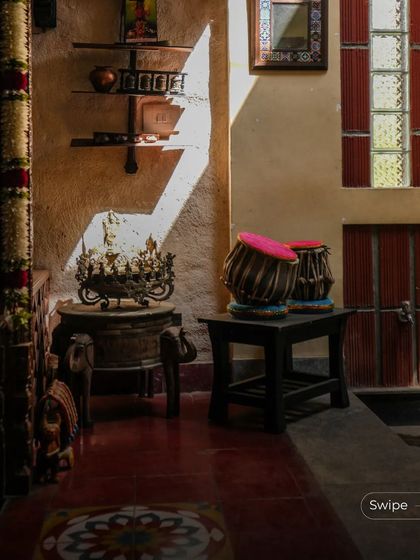 Sunlight streams into a corridor, highlighting traditional decor like a brass urli and a tabla set. The space feels warm, lived-in, and full of cultural resonance.