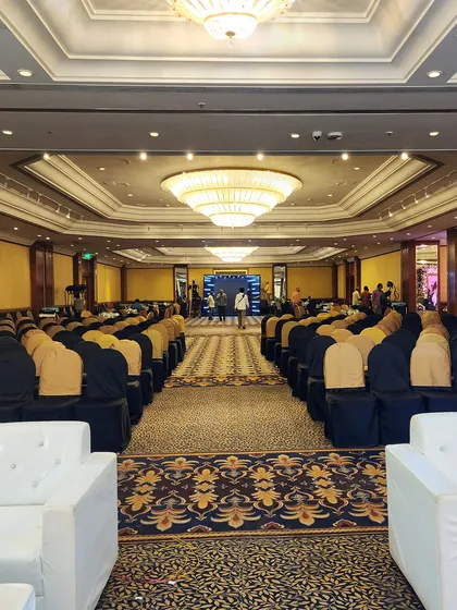 A wide shot of a large ballroom set up for a corporate conference. The seating is arranged theatre-style, facing a stage with a large screen, demonstrating our capability to handle large-scale corporate events.