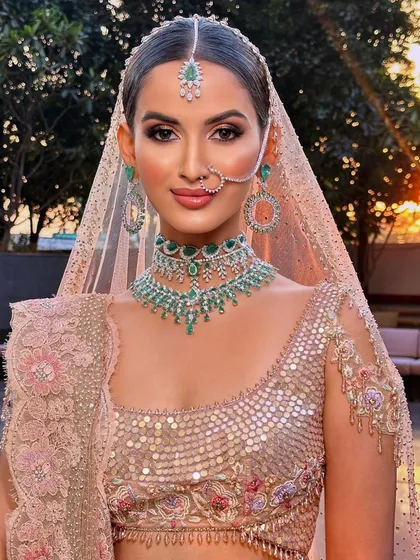 For this modern bridal look, we played with the soft light of the golden hour. The makeup features a dewy, fresh skin finish and soft pink tones on the eyes and cheeks to complement the pastel lehenga and stunning emerald jewellery.