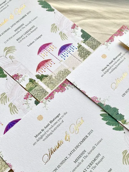 A pattern of Mehendi and Bhaat ceremony cards for the Udaipur wedding. The design combines colorful umbrellas, bougainvillea, and palm illustrations with elegant gold foil script.