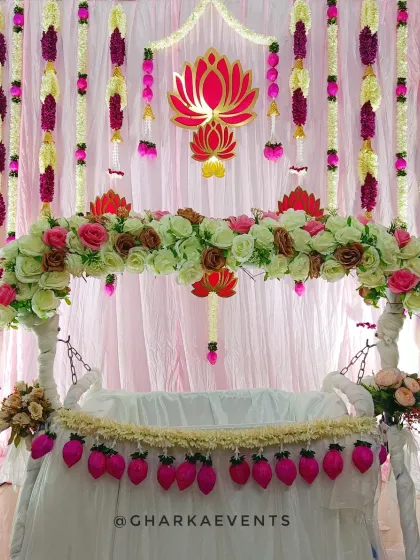 A traditional cradle ceremony decoration. The cradle is adorned with artificial flowers and a lotus motif, set against a backdrop of pink drapes and floral garlands.