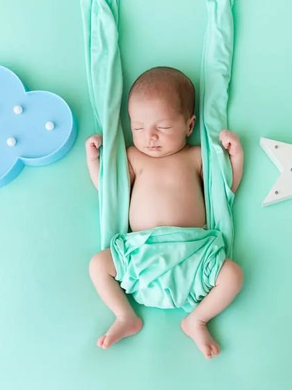 A newborn sleeps peacefully in a mint green sling against a matching backdrop with star and cloud props, creating a dreamy, celestial theme.