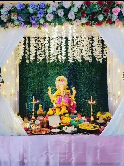This elegant Ganpati mandap features a green grass wall backdrop, draped with white curtains and topped with a colorful floral arrangement. Hanging floral bouquets on the sides complete the look.