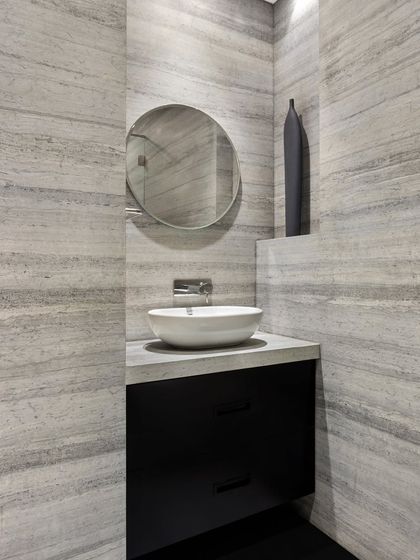 This bathroom in the Limestone Residence uses a serene, grey-veined stone on the walls and countertop. The dark, minimalist vanity cabinet appears to float, creating a sense of lightness in the space.