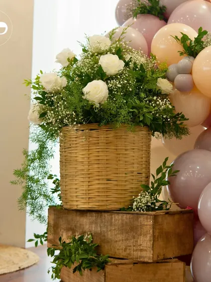Details matter in creating a cohesive look. A rustic wicker basket overflows with fresh white roses and greenery, set against the soft pastel balloon arch. This element adds a natural, garden fresh feel to the indoor picnic theme.