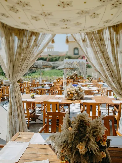 A view from inside a dining cabana, looking out onto the sunlit lawns. The long wooden tables are set with pampas grass centerpieces, creating a cohesive and natural aesthetic.