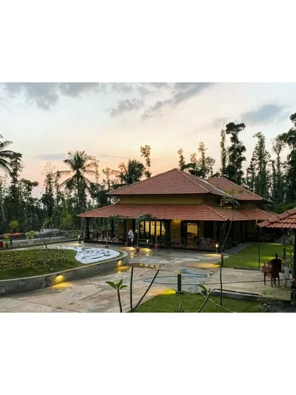 A twilight view of the modernized estate bungalow in Sakleshpur. The warm lighting highlights the traditional tiled roof and veranda, creating an inviting atmosphere amidst the surrounding palm trees and gardens.