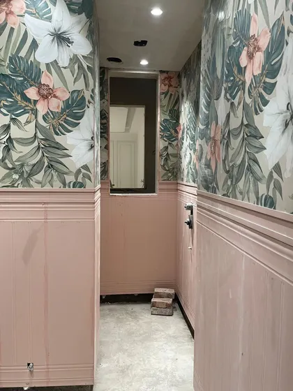 A work-in-progress view of a powder room, showing the installation of floral wallpaper above classic pink wainscoting. This demonstrates the layering of textures and patterns.