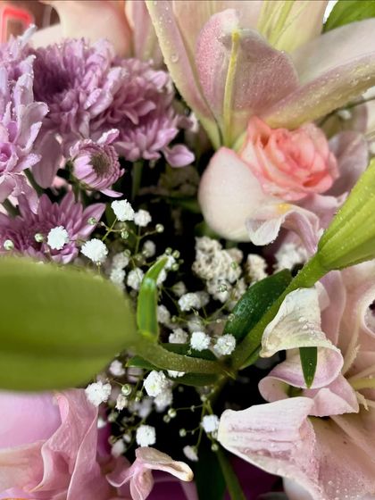 A different angle of the fresh flower arrangement, showing the green leaves and baby's breath that add fullness and contrast to the pink and purple blooms.