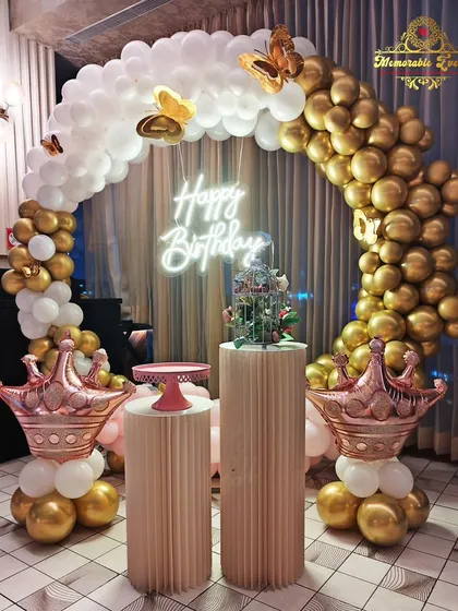 This elegant birthday decoration features a beautiful arch of white and chrome gold balloons, accented with butterfly details and rose gold crown balloons. The setup is completed with stylish plinths and a modern neon 'Happy Birthday' sign.