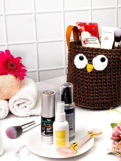 This brown crocheted owl basket is shown in a bathroom setting, holding makeup and skincare products. This demonstrates its versatility as a stylish organizer for any room.
