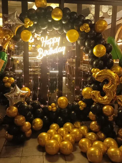 A classy black and gold theme for a 33rd birthday. This ring backdrop is decorated with balloons, a champagne bottle foil, fairy lights, and a neon sign, perfect for a celebration at a restaurant or lounge.