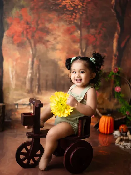 A happy girl on her wooden tricycle, holding a bright yellow flower. The warm, autumnal colors of the backdrop create a cozy and inviting scene.
