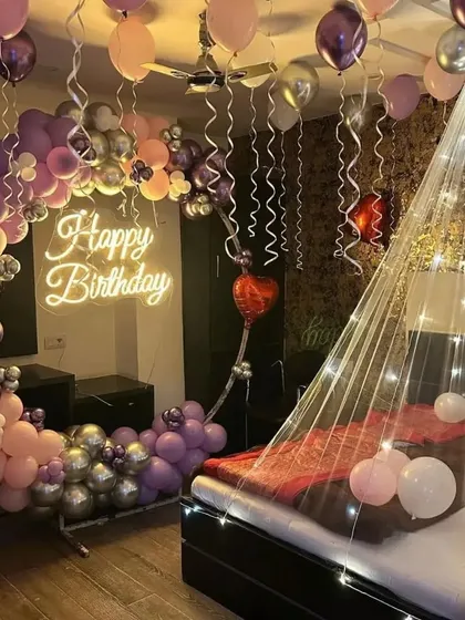 A dreamy birthday room decoration in shades of purple, pink, and silver. The setup includes a crescent-shaped balloon arch next to the bed and a fairy light canopy, creating a magical atmosphere.