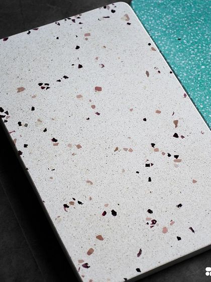 A close-up of a white terrazzo tile with a delicate mix of black and purple chips, next to a vibrant teal sample.