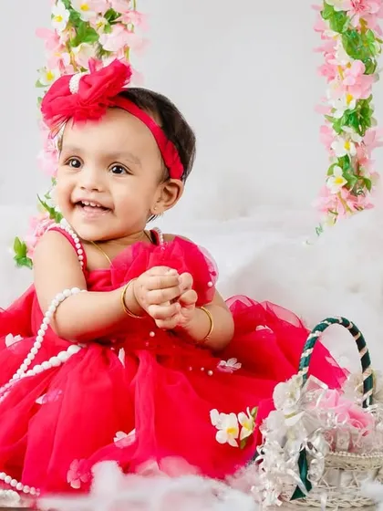 A joyful baby girl in a bright red dress, sitting amidst fluffy clouds and a floral swing for her first birthday.