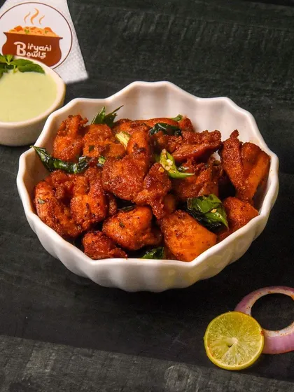 A bowl of spicy Chicken 65, a popular Indian appetizer, garnished with curry leaves and served with mint chutney. The dark background makes the dish look fiery and flavorful.