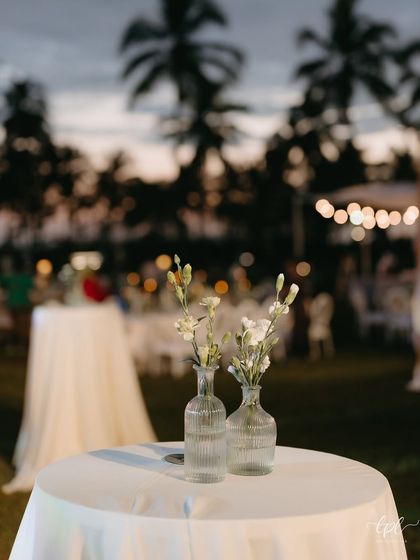 A simple yet elegant detail from a white-themed dinner in Goa. Two small vases with delicate white flowers sit on a cocktail table, with the palm trees and evening sky in the background.