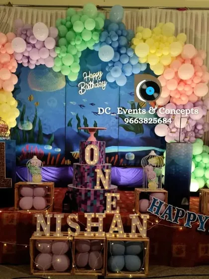An adorable underwater theme for a first birthday party. The backdrop features sea creatures, while a pastel balloon arch and light-up letters spelling 'ONE' complete this charming setup.