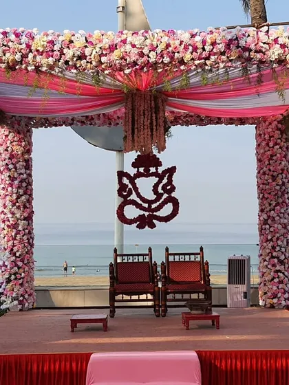 A close-up of a magnificent beachside mandap. The pillars are completely covered in pink flowers, and a floral Ganesh motif provides a traditional centerpiece, all set against the backdrop of the sea.