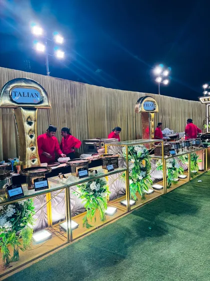 An overview of our elegant Italian food counter at an outdoor evening event. The setup features gold and white decor, beautiful floral arrangements, and dedicated chefs ready to serve fresh pasta and other Italian specialties.