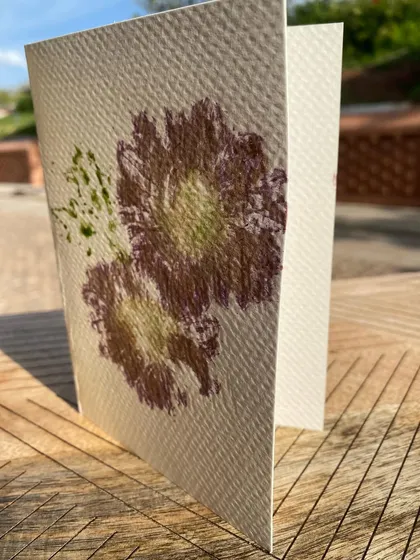 A finished greeting card with a delicate purple flower print, captured in the warm sunlight. These handmade cards are a perfect way to share a piece of nature's beauty.