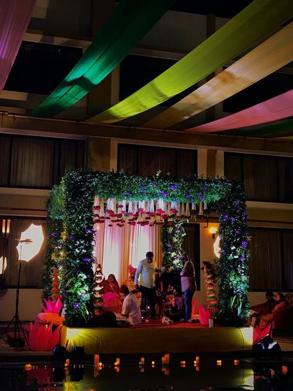 An open-air mandap setup for a night ceremony by the water. The structure is a simple square frame covered in dense greenery and flowers, with colorful drapes overhead, creating a beautiful contrast against the dark sky.