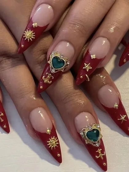 This design shows my skill in creating elaborate, high-fashion nail art. It features long stiletto extensions with a deep red base, adorned with 3D gold charms, sun motifs, and a stunning green heart-shaped jewel.