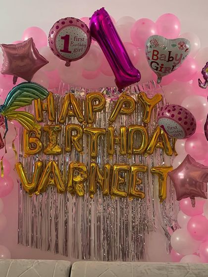 A close-up of the 'Happy Birthday' foil balloon banner with the child's name. This personalized touch is part of a larger pink and white balloon backdrop for a first birthday.