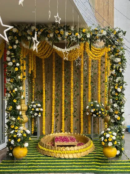 An outdoor haldi setup on a balcony with a beautiful backdrop of marigold strings and white flowers. The decor includes star-shaped lights and a lotus seating area.