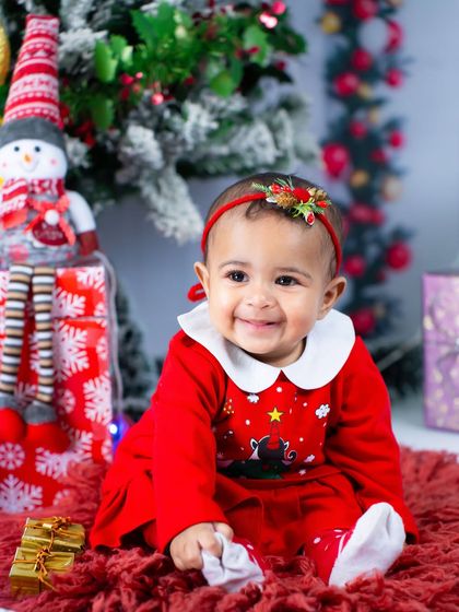 A happy baby girl in her Christmas outfit, surrounded by presents and a festive tree. It's a classic and heartwarming holiday portrait.