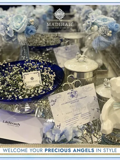 A detailed look at a baby boy announcement hamper, designed to welcome your precious angel in style. The silver tray holds a cobalt blue dish with baby's breath, a crowned teddy bear, and a box of Läderach chocolates.