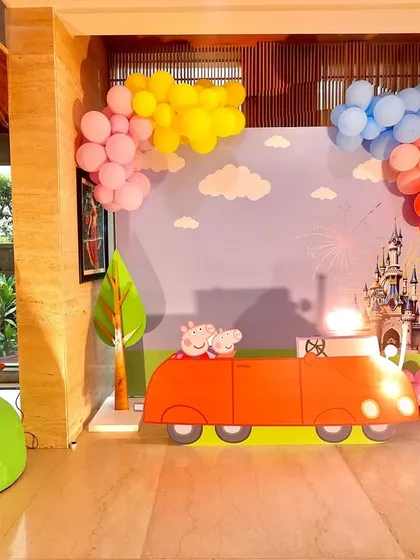 A playful indoor setup for a Peppaland party. The scene includes Peppa's house, her red car against a sky backdrop with a castle, and colorful balloon clouds, creating a multi-layered, imaginative play area.