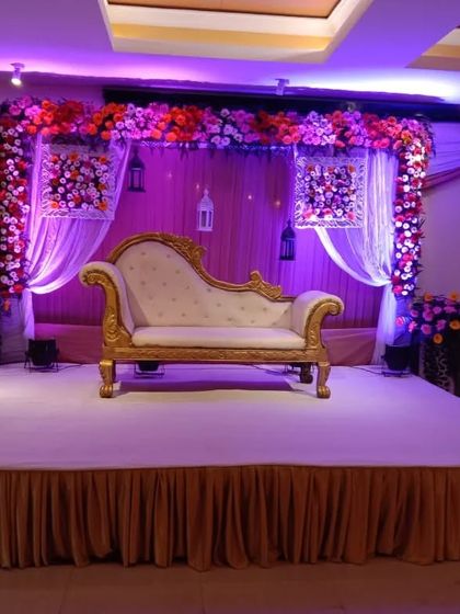 An indoor reception stage with purple uplighting. The decor features a floral arch over a classic white and gold sofa.