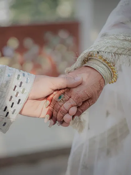 A detailed close-up of the couple holding hands. This shot focuses on the intricate henna on the bride's hand and the beautiful embroidery on their traditional attire, symbolizing their union.