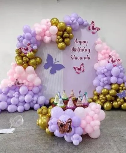 A beautiful butterfly-themed birthday decoration in lavender, pink, and gold. The setup includes a custom backdrop with the name "Shivanchi" and a balloon arrangement perfect for a cake table.