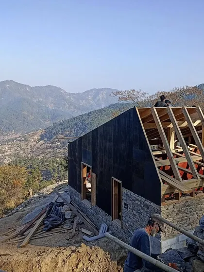 The roof rafters are installed on a guesthouse, with the mountain range visible in the distance. The building is covered with a protective layer, shielding the stonework from the elements during construction.