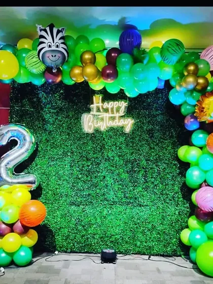 For a kid's second birthday, I made this jungle theme decoration. The setup features a green grass wall, a balloon arch with animal foils, a 'Happy Birthday' neon sign, and a number '2' balloon pillar.
