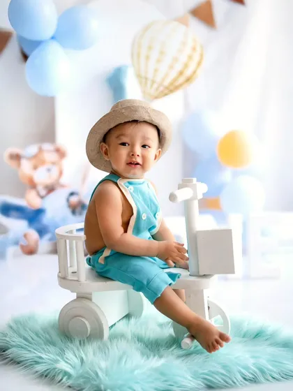 A happy little traveler on his first birthday adventure. Dressed in a cute hat and overalls, he's ready to explore from his little white toy bike.