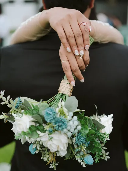 A bride's hands, adorned with beautiful nails and rings, holding her bouquet from behind her groom. This detailed shot from Anu and Nikhil's wedding is a beautiful symbol of their union.