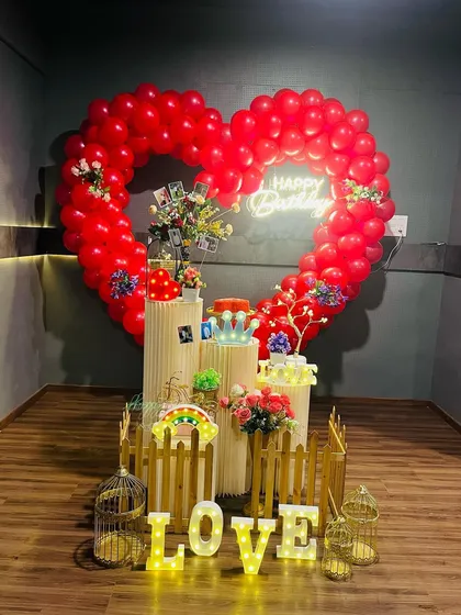 A romantic surprise birthday decoration with a large heart-shaped balloon frame. The setup is filled with personal touches, including photos, light-up "LOVE" letters, and a neon "Happy Birthday" sign.