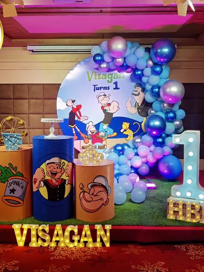A classic Popeye the Sailor-themed first birthday. The setup features character-printed plinths, a blue and silver balloon arch, and a 'VISAGAN' marquee.