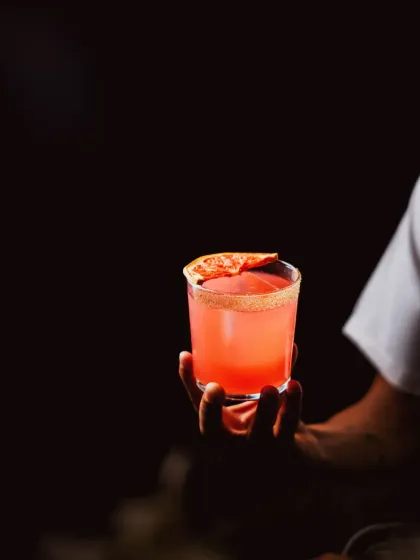 A hand holds our Pimento Paloma, a bold blend of tequila, grapefruit, and passion fruit, with a touch of litchi to soften the jalapeño heat.