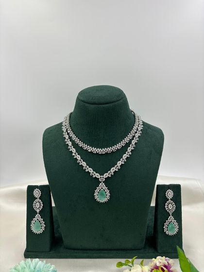 A graceful double-layered AD necklace. The top layer is a simple floral chain, while the bottom layer features a pear-shaped pendant. It is available in white, pink, green, and blue stones.
