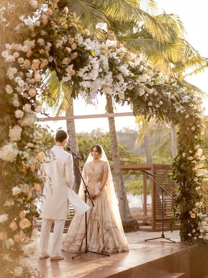 Vows exchanged under a beautiful floral arch. The bride's ethereal ivory Tarun Tahiliani lehenga and the groom's matching classic sherwani create a picture of pure elegance against the beach backdrop.