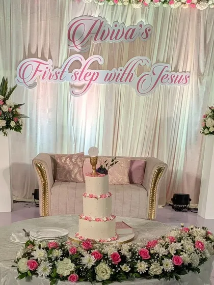 The cake table and stage backdrop for Aviva's 'First step with Jesus.' The design features angelic cutouts, soft draping, and beautiful floral arrangements in a pastel pink and white palette.