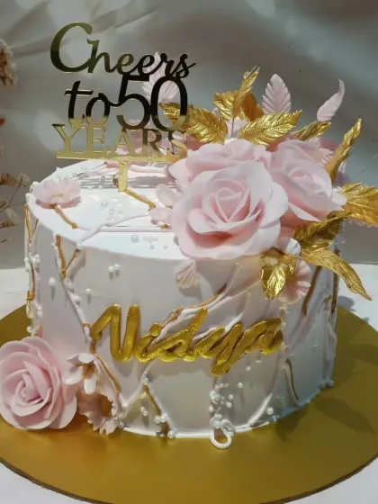 A beautiful cake for a 50th birthday, featuring soft pink sugar roses, delicate white piping, and artistic gold-painted leaves. The personalized name in gold script adds a final touch of elegance.
