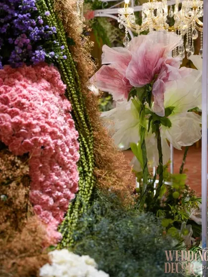 A close-up of the floral gown sculpture, showing the intricate layering of different flowers and textures. The giant paper flowers in the background add to the whimsical, fairy-tale feel.